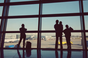 Four Sihloutted People Standing Looking Out Of An Airport Window Onto The Runway