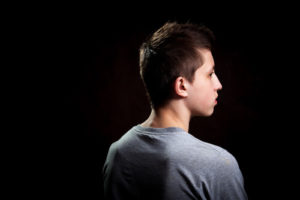 White Male Teen In Grey T-Shirt With Back Turned Away In Front Of A Black Background