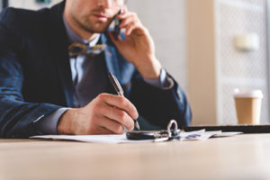 White Male In A Blue Suit Sitting At A Table While Talking On A Cell Phone And Jotting Down Notes
