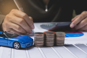 Die-Cast Blue Corvette And Several Stacks Of Coins On Table In front Of Man Holding Pen Using A Calculator