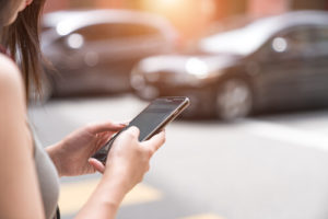 White Woman Standing In Front Of Two Silver Sedans Holding A Smart Phone