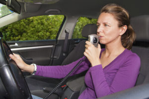 White Woman In Purple Longsleeve Shirt Siting In Drivers Seat Breathing Into Breathalyzer