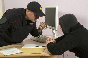 Police Officer Holding Handcuffs While Menacingly Leaning Over Desk Towards A Seated White Juvenile Male